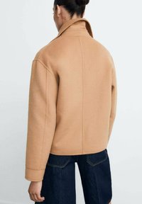 Persoon met een bun in het haar, gekleed in een camelkleurige wollen jas en donkere denim broek, gezien van achteren tegen een effen achtergrond.