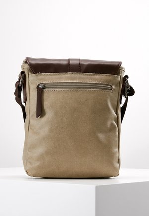 Beżowa torba na ramię z płótna z brązową skórzaną klapką, przednią kieszenią na zamek i regulowanym paskiem na ramię na białym tle.