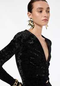 Frau trägt ein strukturiertes schwarzes Kleid mit tiefem V-Ausschnitt, große goldene Ohrringe in Form von Blättern und dicke goldene Armbänder, vor weißem Hintergrund.