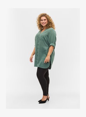 Groene linnen button-up tuniek met opgerolde mouwen, gecombineerd met zwarte leggings met knoopaccenten aan de zoom. Model draagt zwarte hakken.