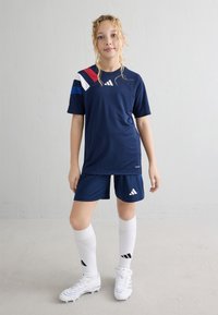 Fotbalový dres Adidas v námořnické modři, který obsahuje tričko s krátkým rukávem s červenými, bílými a modrými detaily, odpovídající šortky, bílé ponožky a kopačky.
