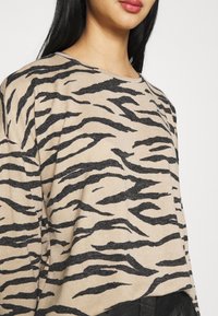 Beiger Pullover mit einem schwarzen Tigerstreifenmuster, langen Ärmeln und einem lockeren Schnitt. Weicher Stoff mit weiter Halsöffnung. Schwarze Akzente am Saum.