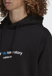 Sort hættetrøje lavet af stof med en blød tekstur. Har et frontlogo i hvidt og blåt, med snoretræk i hætten og ribbede detaljer.