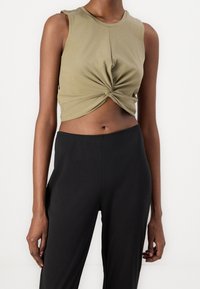 Oliwkowy bezrękawnik w skróconym fasonie z zawiązanym przodem, zestawiony z czarnymi spodniami o wysokim stanie. Miękka tkanina o swobodnym kroju.