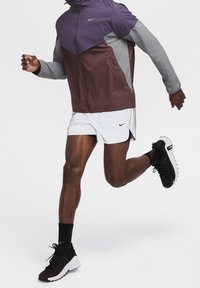Läufer in einer farbigen Windjacke, weißen Shorts, schwarzen Socken und schwarz-weißen Laufschuhen inmitten eines Schrittes vor einem schlichten Hintergrund.
