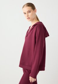 Burgundowy hoodie z dekoltem w serek i luźnym krojem. Wykonany z miękkiego materiału, z kapturem i długimi rękawami. Widoczne szczegóły przeszyć.