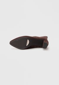 Brown suede high-heeled shoe with a tapered toe and a textured rubber sole. Features a small block heel and branded accent on the sole.