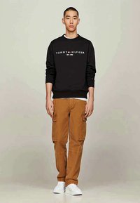 Hanorac negru cu logo alb "TOMMY HILFIGER"; pantaloni cargo bej cu buzunare laterale; adidași albi; fundal neutru.
