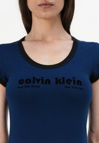 Tamsiai mėlynas medvilninis marškinėlis su juodu apvaliu kaklu. Yra ryškus juodas logotipo tekstas su užrašu "calvin klein" ir vietos informacija po juo.