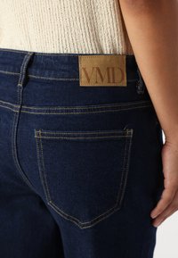 Tamno plave traperice s zlatim šavovima, kožnom oznakom s natpisom "VMD" i klasičnim dizajnom stražnjih džepova. Prikaz blizu pojasnice.