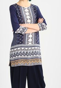 Marinblå och vit mönstrad tunika med trekvartslånga ärmar, som har texturerade detaljer, intrikata mönster och en delad fåll.