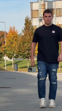 Juoda trumpomis rankovėmis marškinėliai, mėlyni su dėvėjimo žymėmis džinsai ir balti sportbačiai su smėlio spalvos akcentais. Fone matyti rudens medžiai ir šaligatvis.