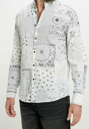 Mann trägt ein weißes langärmliges Hemd mit schwarzen, aufwendigen Paisley- und Patchworkmustern, kombiniert mit dunklen Jeans.