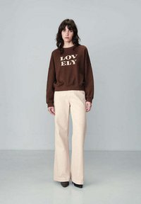 Felpa marrone con la scritta "LOVELY" stampata in beige, abbinata a pantaloni beige a gamba larga. L'outfit presenta una vestibilità morbida e un tessuto testurizzato.