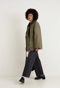 Jachetă oversized, ușoară, de culoare verde măslin, cu mâneci lungi și buzunare în față, asortată cu blugi negri cu picioare largi și pantofi negri.