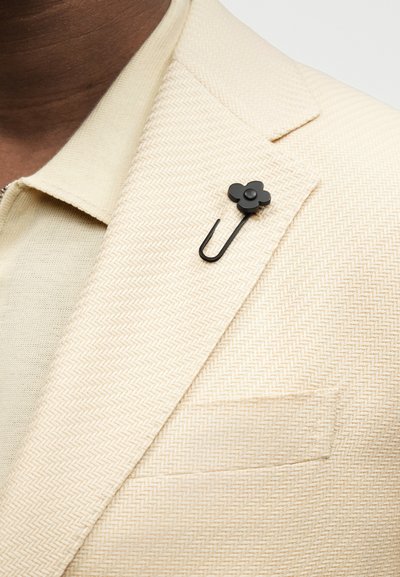 Μαύρο καρφί τριγωνικού σχήματος σε λουλούδι, τοποθετημένο στο αριστερό πέτο ενός μπεζ σακακιού με σχέδιο ψαροκόκκαλο, που φοριέται πάνω από ένα μπεζ πλεκτό πουκάμισο.