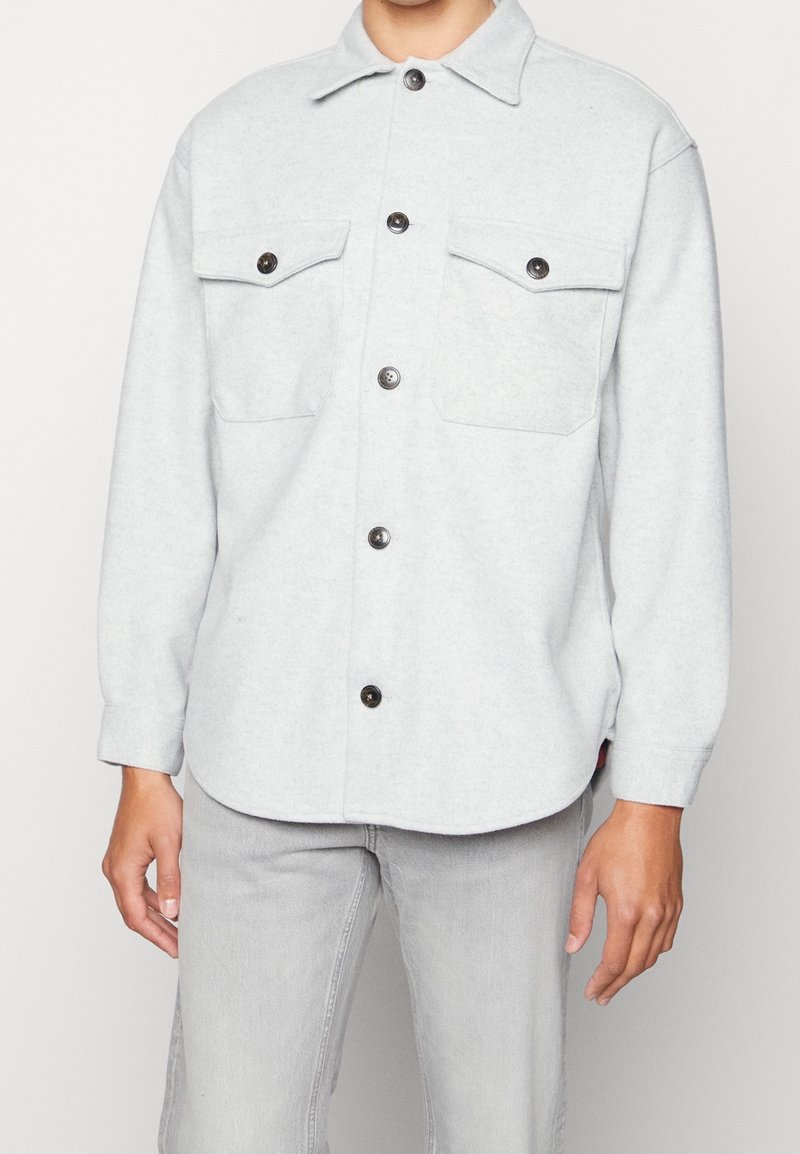 Lys grå flannel skjorte med lange ærmer, knappetryk fortil, to brystlommer og sorte knapper. Blød tekstur og rundet kantdesign.