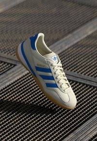 Crème sneakers met blauwe strepen en gouden "Gazelle" tekst. Beschikt over een gestructureerde zool en een klassiek low-top ontwerp.