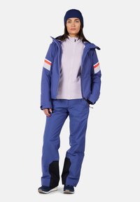 Giacca impermeabile blu con strisce rosse e bianche sulle maniche, abbinata a pantaloni blu coordinati e un berretto di lana blu navy. Tessuto testurizzato in tutto.