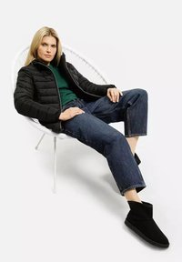 Czarna pikowana kurtka, ciemnozielony sweter, luźne ciemnoniebieskie dżinsy i czarne botki za kostkę, siedząca w okrągłym białym fotelu.