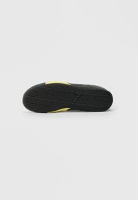 Must kummikute tald detailse kulumisprofili ja Puma logo, kollased küljed selgelt nähtavad lihtsa valge tausta taustal.