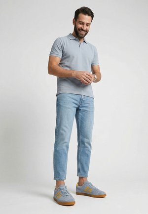 Uśmiechnięty mężczyzna poprawiający zegarek na rękę, ubrany w jasnoszary polo, jasnoniebieskie dżinsy i szare trampki z żółtymi paskami, stojący na jednolitym tle.