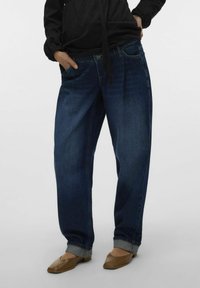 Denim hlače v temno modrem odtenku, z udobnim krojem, zavihanimi robovi in sprednjimi žepi. V kombinaciji s črnim zgornjim delom in ravnimi čevlji.