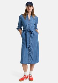 Denim krekli kleita ar garām piedurknēm, pogājamu priekšpusi, sašutu jostu un sānu kabatām. Apvienota ar sarkanām sporta apavām un baltām zeķēm.