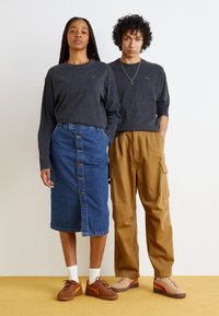 Două modele stau unul lângă celălalt. Primul poartă o cămașă cu mânecă lungă de culoare cărbune și o fustă din denim tip creion cu nasturi. Al doilea, îmbrăcat într-o cămașă asortată, are pantaloni cargo khaki. Amândoi poartă încălțăminte casual în nuanțe pământii.