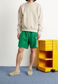 Beige sweatshirt med rund halsringning, tillsammans med gröna träningsshorts med vita sidostreck och en logotyp. Ljusgrå skor. Gul kundvagn i närheten.