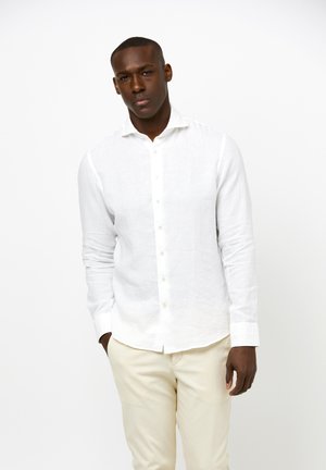 Man wearing a white button-up long-sleeve shirt and beige pants standing against a plain white background.