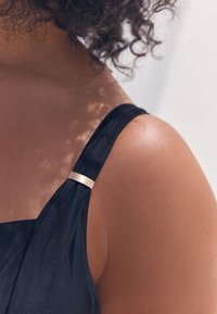 Indumento nero con una texture liscia e una cinghia larga, con un dettaglio in metallo dorato vicino alla spalla. Tono della pelle visibile sotto.