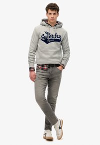 Felpa grigia con logo blu navy, camicia a quadri sotto, jeans grigi e sneakers bianche con suole color sabbia. Presenta un taglio casual e tasca frontale.