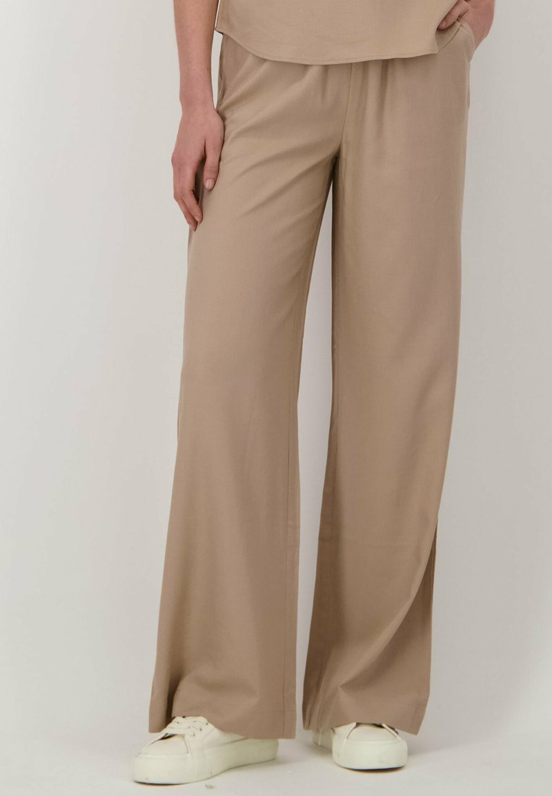 Beige wijd uitlopende broek van lichtgewicht stof, met een ontspannen pasvorm, elastische tailleband en zijzakken. Gepaarde met witte sneakers.