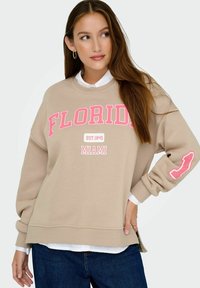 Sweatshirt marron clair avec le texte "Florida" en rose et "Miami" en dessous. Manches longues et ourlet côtelé, superposé sur une chemise blanche à col.