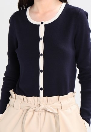 Kobieta ubrana w granatowy kardigan z długim rękawem, ozdobiony białym obszyciem i guzikami, w zestawie z beżowymi spodniami z wysokim stanem typu paperbag, związanymi w talii.