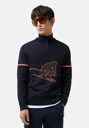 Marineblauer Strickpullover mit halbem Reißverschlusskragen; verfügt über eine orange Skifahrer-Grafik und einen horizontalen Streifen; kombiniert mit übergroßen Sonnenbrillen.