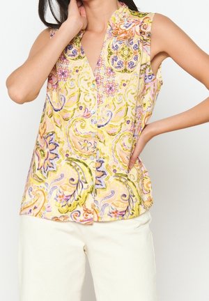 Vrouw die een mouwloze, geknoopte blouse draagt met meerkleurige bloemen- en paisleyprint, gecombineerd met lichtgekleurde broek, poseert met één hand op de heup.