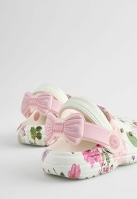 Zoccoli a slip-on floreali con base bianca, dettagli a fiocco rosa e suola testurizzata. Presenta un mix di motivi floreali rosa e verdi.