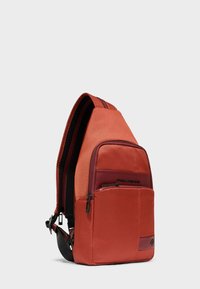 Rode schouderrugzak met voorvak met rits en verstelbare zwarte riem, rechtopstaand afgebeeld tegen een effen lichtgrijze achtergrond.