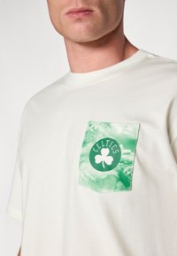 Bijela pamučna majica s grafičkim džepom u zelenoj boji koji sadrži djetelinu i tekst "CELTICS", okrugli ovratnik i kratke rukave.