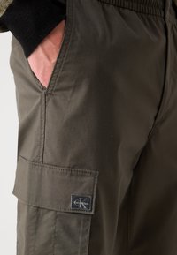 Olive zelené cargo nohavice vyrobené z odolnej tkaniny, so bočnou vreckom s čiernou nášivkou loga Calvin Klein a elastickým pásom.