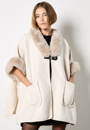Jonge vrouw die een crèmewitte wollen cape met beige nepbont trim aan de kraag en mouwen draagt, met handen in de voorzakken tegen een effen achtergrond.