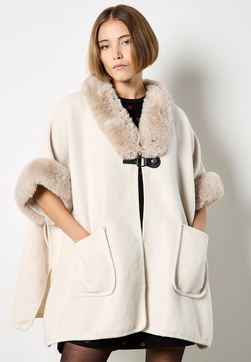 Jonge vrouw die een crèmewitte wollen cape met beige nepbont trim aan de kraag en mouwen draagt, met handen in de voorzakken tegen een effen achtergrond.