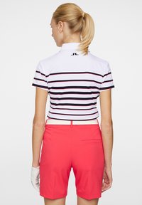 Vit skjorta med marinblå och röda ränder, kombinerad med klara röda shorts. Inkluderar en bakficka och ett vitt bälte. Tillverkad av syntetiskt material.