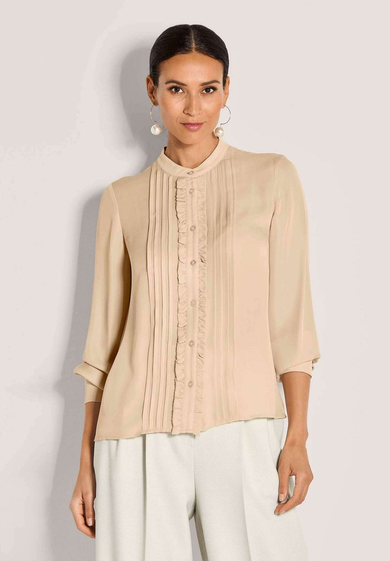 Beige Bluse mit gerüschtem Vorderteil, Knopfverschluss und 3/4-Ärmeln. Hergestellt aus glattem Stoff, der vertikale Falten für Struktur aufweist.