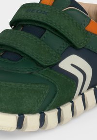 Gros plan sur une sneaker verte et bleu marine avec deux bandes Velcro, semelle blanche, détail orange et une main tenant la chaussure.