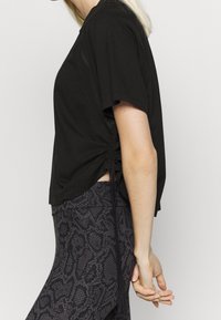 Kvinna klädd i en svart kortärmad topp med rynkade sidor och högmidjade leggings med svart och grå mönster, stående på sidan.