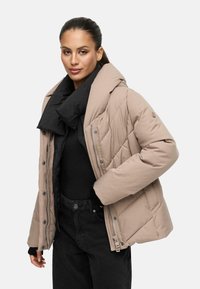 Veste rembourrée beige avec une écharpe noire amovible. Elle comporte une capuche, une fermeture zippée et des textures contrastées provenant du design matelassé.