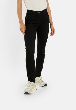 Persona con vaqueros negros ajustados y zapatillas beige y plateadas, de pie frente a un fondo claro y liso.